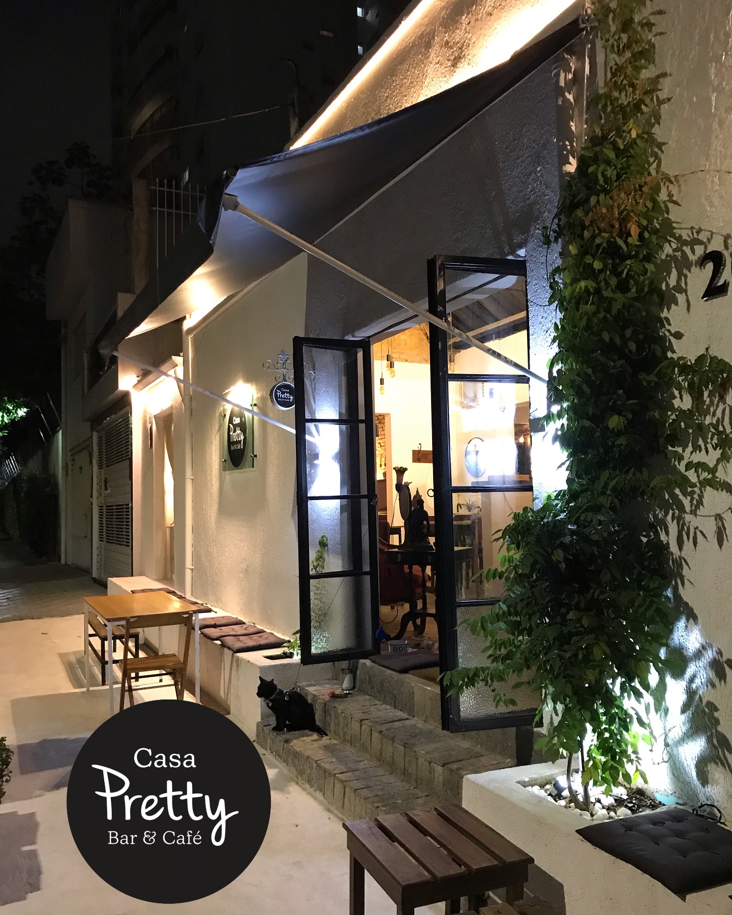 Fachada da Casa Pretty Bar & Café à noite, com toldo iluminado e mesas na calçada